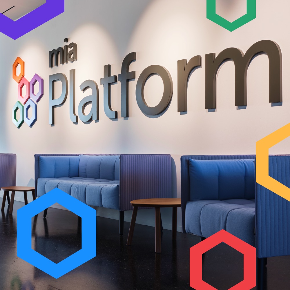 Parete della reception Mia‑Platform con il logo aziendale 3D, divani blu e esagoni colorati sovrapposti alla foto